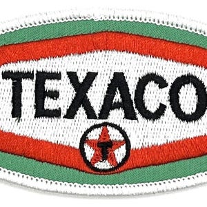 May include: An oval-shaped embroidered patch with a white background and red border. The text "TEXACO" is in black lettering. A red star with a black outline and a black "T" inside is in the center of the patch. The patch is surrounded by a white border.