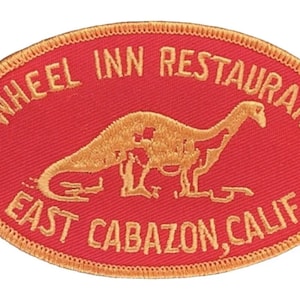 Puede incluir: Un parche bordado de forma ovalada roja con costuras doradas. El parche presenta la silueta de un dinosaurio y el texto "WHEEL INN RESTAURANT EAST CABAZON, CALIF."