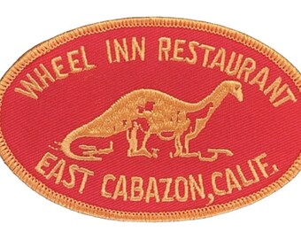 May include: A red oval-shaped embroidered patch with gold stitching. The patch features a dinosaur silhouette and the text "WHEEL INN RESTAURANT EAST CABAZON, CALIF."