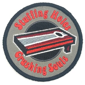 May include: A gray and black embroidered patch with red stitching. The patch features a red, white, and gray cornhole board with the text "Stuffing Holes Crushing Souls".