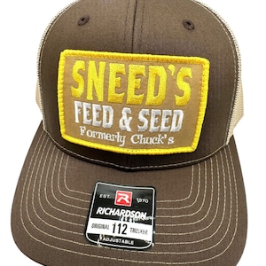 Może przedstawiać: Brązowa i beżowa czapka trucker z żółto-brązową naszywką z napisem "SNEED'S FEED & SEED Formerly Chuck's". Czapka ma czarną i czerwoną metkę z napisem "EST. R 1970 RICHARDSON ORIGINAL 112 TRUCKER ADJUSTABLE".