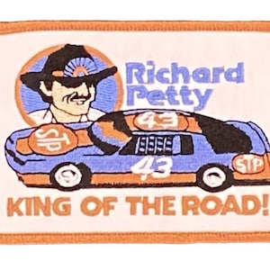 May include: An embroidered patch featuring a cartoon image of Richard Petty, a race car driver, wearing a hat and driving a blue race car with the number 43 on the side. The car is sponsored by STP. The text "Richard Petty" and "King of the Road!" are also on the patch.