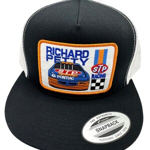 May include: Black and white trucker hat with a black and orange patch featuring a race car and the text "Richard Petty STP Racing". The hat has a white mesh back and a black snap closure with the text "The Authentic Yupoong Snapback Yupoong Since 1974".