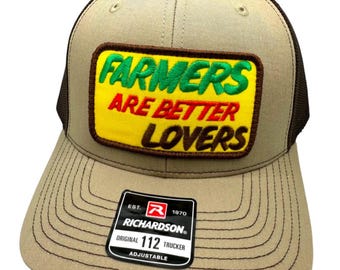 May include: A brown and black trucker hat with a yellow, green, and red patch that says "Farmers Are Better Lovers". The hat has a black mesh back and a brown front with a black Richardson 112 label.