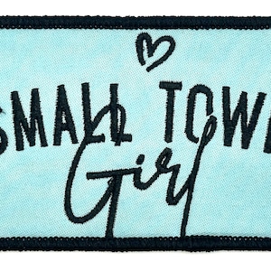 Op de afbeelding: Een rechthoekige patch met een lichtblauwe achtergrond en een zwarte rand. De patch bevat de woorden "SMALL TOWN Girl" in zwart schrift en een klein hartje boven het woord "TOWN".