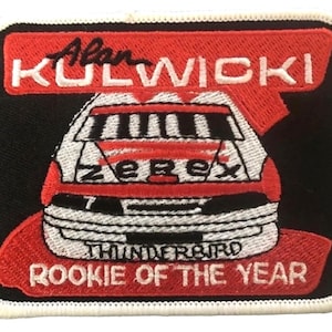 May include: A red, white, and black embroidered patch with the text "Alan Kulwicki" and "Rookie of the Year". The patch features a white race car with a red stripe and the word "Zerex" on the side. The car is a Thunderbird.