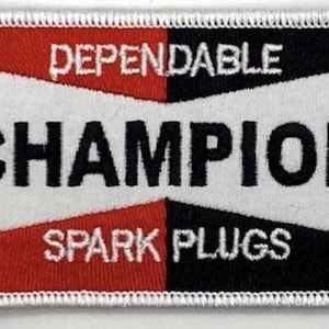 Könnte beinhalten: Rechteckiger, gestickter Aufnäher mit dem Text "CHAMPION SPARK PLUGS". Das Design hat einen weißen Rand, einen roten und schwarzen Hintergrund, weiße geometrische Formen und das Wort "CHAMPION" in fetten schwarzen Buchstaben.