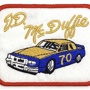 Può includere: Toppa ricamata con un'auto da corsa blu e oro con il numero "70" sul lato. Il testo "J.D. McDuffie" è ricamato in oro sopra l'auto.