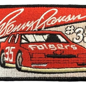 Pode incluir: Etiqueta bordada vermelha e branca com um carro de corrida vermelho com o número 35 no lado e as palavras "Folgers" na frente. A etiqueta também inclui a assinatura "Kenny Irwin" e o número "#35" em branco.