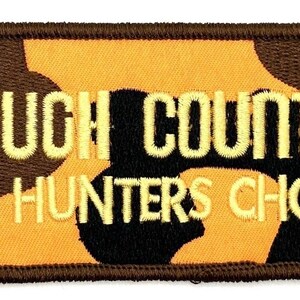 May include: Embroidered patch with the text "ROUGH COUNTRY the HUNTERS CHOICE". The patch has a brown border and a camouflage pattern in shades of brown, black, and yellow. The text is in a yellow, stitched font.