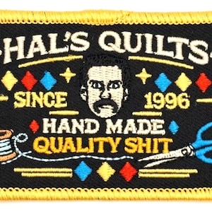 Puede incluir: Parche bordado con fondo negro y borde amarillo. El parche presenta el texto "HAL'S QUILTS" sobre una cara estilizada con bigote. Debajo están las palabras "HAND MADE QUALITY SHIT" y el año "1996".