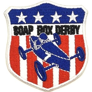 May include: A white, red, and blue embroidered patch with a soapbox derby car in the center. The patch has a shield shape with three white stars in the top blue section. The text "SOAP BOX DERBY" is in black letters above the car.