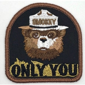 Könnte beinhalten: Brauner gestickter Aufnäher mit einem Cartoon von Smokey Bear, der einen Rangerhut mit dem Wort "SMOKEY" trägt. Der Aufnäher hat einen braunen Rand und die Worte "ONLY YOU" in braunen Buchstaben auf einem beigefarbenen Hintergrund.