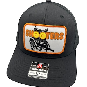 May include: Black trucker hat with a white patch featuring a cartoon owl holding a rifle and the text "SHOOTERS". The patch has an orange border. The hat has a black mesh back and a black adjustable strap with the text "Richardson 112 Trucker Adjustable".