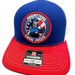 May include: A blue and red Richardson 112 trucker hat with a white mesh back. The hat features a circular patch with the words "PIPEFITTERS UNION PROUD" and an image of a pipefitter. The brim is red.