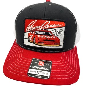 Pode incluir: Boné de caminhoneiro preto e branco com um remendo vermelho e branco que mostra um carro de corrida vermelho com o texto "Benny Parsons #35 Folgers". O boné tem uma aba vermelha com costuras brancas e uma parte de trás de malha preta e branca. O boné tem um logotipo Richardson preto e vermelho com o texto "EST. 1970 RICHARDSON® ORIGINAL 112 TRUCKER ONE SIZE".