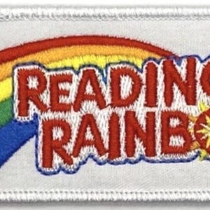May include: Embroidered patch with a rainbow and the words "READING RAINBOW" in red, with a blue outline. The patch has a white background and a white stitched border. A yellow sunburst design is next to the word "RAINBOW".