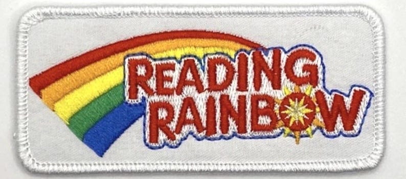 K&ouml;nnte beinhalten: Gestickter Aufn&auml;her mit einem Regenbogen und den Worten "READING RAINBOW" in Rot, umrandet in Blau. Der Aufn&auml;her hat einen wei&szlig;en Hintergrund und einen wei&szlig;en, gen&auml;hten Rand. Ein gelbes Sonnenstrahl-Design befindet sich neben dem Wort "RAINBOW".