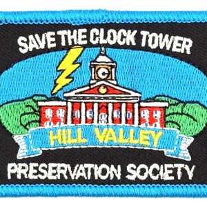 May include: Embroidered patch with a black background and a blue border. The patch features a clock tower illustration with the text "SAVE THE CLOCK TOWER" above it. Below the tower is a banner that reads "HILL VALLEY". The text "PRESERVATION SOCIETY" is below the banner.