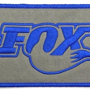Könnte beinhalten: Ein blauer gestickter Aufnäher mit dem Wort "FOX" in einer stilisierten Schriftart. Der Aufnäher befindet sich auf einem grauen Hintergrund und hat einen blauen Rand.