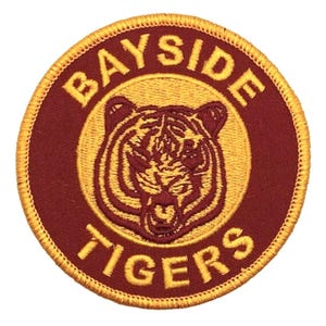 May include: A circular patch with a maroon background and gold embroidered details. The patch features the text "BAYSIDE TIGERS" in a gold, arched font around a gold tiger head.