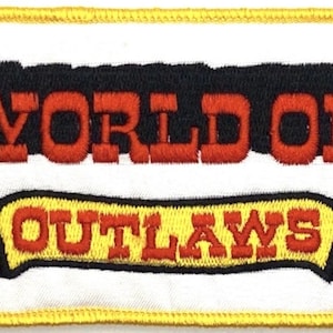 Pode incluir: Patch bordado com as palavras "WORLD OF OUTLAWS" em letras vermelhas. O texto está em um fundo branco com uma borda preta e amarela. O patch é retangular com uma borda costurada amarela.