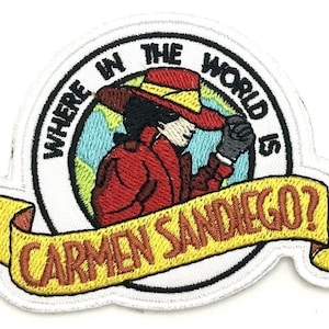 May include: Embroidered patch featuring a cartoon image of Carmen Sandiego in a red coat and hat, with the text "WHERE IN THE WORLD IS CARMEN SANDIEGO?" in a circular design and banner.
