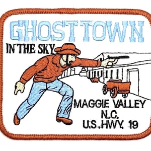 May include: Embroidered patch with a brown border and a white background. The patch features a cowboy shooting a gun, with the text "GHOST TOWN IN THE SKY" in light blue. Below the cowboy is the text "MAGGIE VALLEY N.C. U.S. HWY. 19".