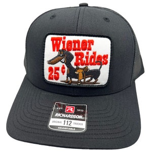 May include: Black trucker hat with a red and white patch featuring a cartoon dachshund with a saddle and the text "Wiener Rides 25¢". The hat has a black mesh back and a black adjustable strap with the text "Richardson 112 Original Trucker Adjustable".
