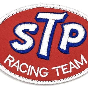 May include: Oval-shaped STP Racing Team patch. The patch features the STP logo in white with blue trim, set against a red background. The words "RACING TEAM" are embroidered in white along the bottom edge.
