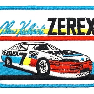 May include: A white race car with a blue, red, and yellow stripe design. The car has the number 7 on the side and the word "Zerex" on the front. The car is on a blue background with the words "Alan Kulwicki Zerex" above it.
