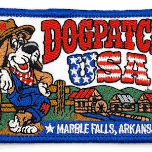 May include: A rectangular embroidered patch with a blue border featuring a cartoon dog in a hat and blue overalls leaning on a fence. The text reads DOGPATCH USA in red and white, with MARBLE FALLS, ARKANSAS at the bottom over a rural landscape.