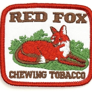 Puede incluir: Parche bordado con un zorro rojo acostado en la vegetación verde. El texto "Red Fox" está en letras marrones sobre el zorro y "Chewing Tobacco" está en letras marrones debajo del zorro.