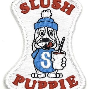 Peut inclure: Patch brodé blanc avec un chien de dessin animé portant un chapeau bleu et une chemise bleue avec un "S" blanc dessus. Le chien tient une tasse blanche avec une boisson rouge et une paille. Le texte "Slush Pupie" est en rouge au-dessus et en dessous du chien.