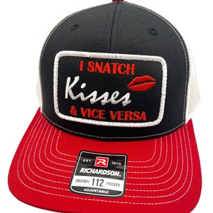 May include: A black and white trucker hat with a red bill. The hat has a white patch with red embroidery that says "I snatch kisses & vice versa". The hat is adjustable and has a Richardson logo on the side.