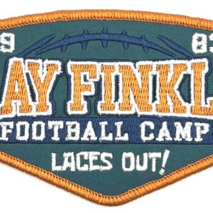 Puede incluir: Un parche bordado de color verde y naranja con el texto "Ray Finkle Football Camp Laces Out!" y los números "19" y "83".