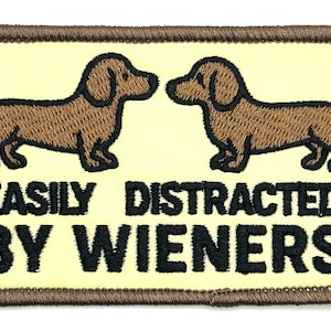 May include: Embroidered patch with two brown dachshunds facing each other. The patch has a light yellow background with the text "EASILY DISTRACTED BY WIENERS" in black, and a brown border.