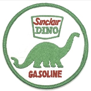 Puede incluir: Parche bordado con el logotipo de Sinclair Oil, un dinosaurio verde y las palabras "Sinclair DINO GASOLINE" en rojo y verde. El parche tiene un fondo blanco con un borde verde cosido.