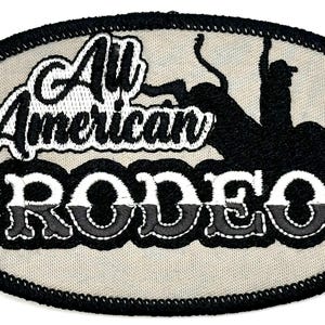 Op de afbeelding: Ovale patch met de tekst "All American Rodeo" in witte en zwarte stiksels. Een silhouet van een rodeo-ruiter op een bokstier is ook aanwezig. De patch heeft een beige achtergrond en een zwarte rand.