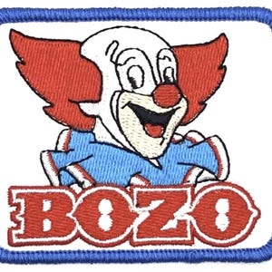 Puede incluir: Parche bordado con un payaso sonriente con pelo y nariz rojos, vestido con un atuendo blanco y azul. La palabra "BOZO" se muestra en rojo, delineada en blanco, en la parte inferior del parche. El parche tiene un borde azul.