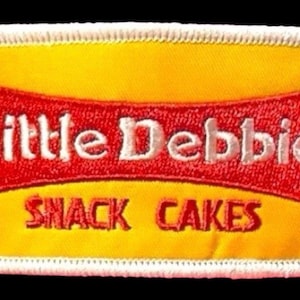 May include: A rectangular embroidered patch with a yellow background and a white stitched border. It features a red horizontal banner containing the white text Little Debbie, with the red text SNACK CAKES printed directly below the banner.