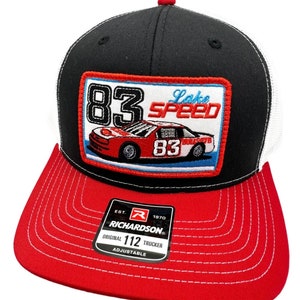 May include: Black and white trucker hat with a red and blue embroidered patch featuring a race car with the number 83 and the text "Lake Speed 83". The hat has a red brim and a white mesh back. The hat also has a black label with the text "Richardson 112 Trucker Adjustable" and a red "R" logo.