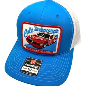 Puede incluir: Un gorro de camionero azul y blanco con un parche bordado en rojo, blanco y azul que presenta un coche de carreras y el texto "Dale Yarborough Hardee's Hardee's Racing". El gorro tiene una etiqueta de camionero Richardson 112.