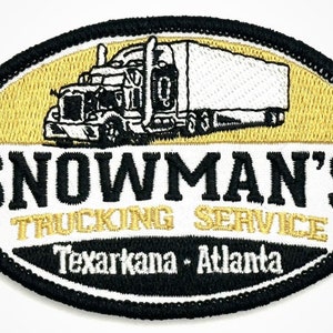 May include: A white oval patch with black trim. The patch features a black and white illustration of a semi-truck with a trailer. The text "SNOWMAN'S TRUCKING SERVICE Texarkana - Atlanta" is embroidered in black and gold.