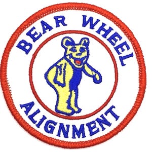 May include: A white embroidered patch with a red border. The patch features a yellow bear with blue arms and legs, standing on its hind legs. The text "BEAR WHEEL ALIGNMENT" is written in a blue circle around the bear.
