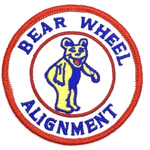 May include: Embroidered patch featuring a yellow and blue cartoon bear with a red mouth. The patch has a white background with the words "BEAR WHEEL ALIGNMENT" in blue, encircled by a red border.