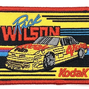 Könnte beinhalten: Gelber und schwarzer gestickter Aufnäher mit einem Rennwagen mit dem Text "Rick Wilson" und "Kodak".