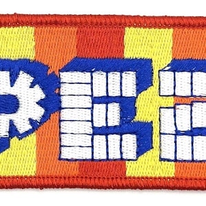 以下が含まれることがあります： 白、青、黄色の「PEZ」の文字が刺繍されたパッチ。文字はブロック状で様式化されており、特徴的な青色の「P」のデザインがあります。背景は赤、オレンジ、黄色の縦縞で、赤い縁取りがあります。