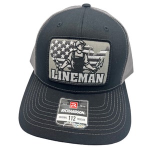 Può includere: Cappellino da camionista nero e grigio con una toppa che raffigura un elettricista che tiene dei fili davanti a una bandiera americana. La toppa riporta la scritta "Lineman". Il cappello ha una visiera nera e una rete grigia sul retro. Il cappello ha un'etichetta con la scritta "Richardson" e "Original 112 Trucker".