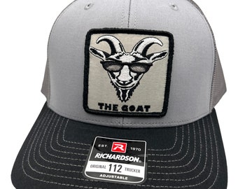 May include: A gray and black trucker hat with a white patch featuring a goat wearing sunglasses and the text "THE GOAT". The hat has a black adjustable strap with a white label that reads "EST. 1970 RICHARDSON ORIGINAL 112 TRUCKER ADJUSTABLE".